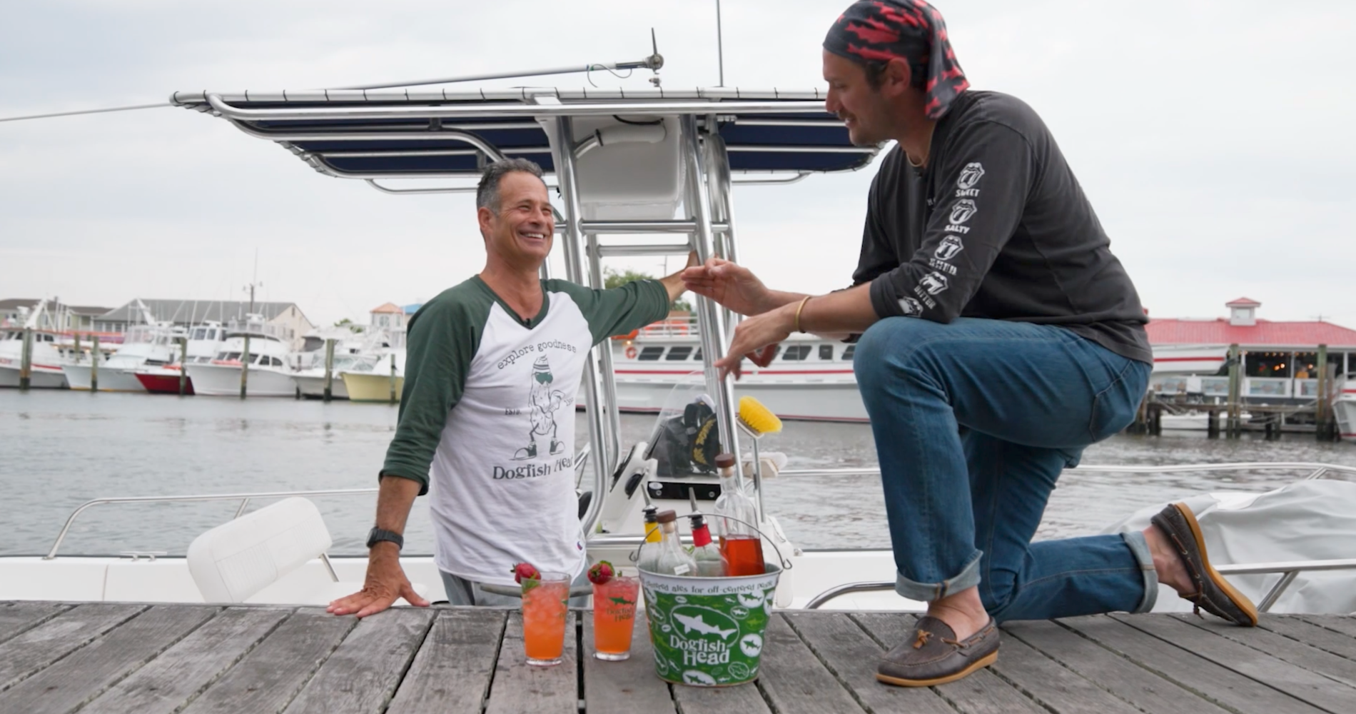 Sam Calagione and brad Leone with two pink cocktails in hurricane glasses on a dock in Lewes, Delaware with the S.S. Dogfish boat in the water behind them