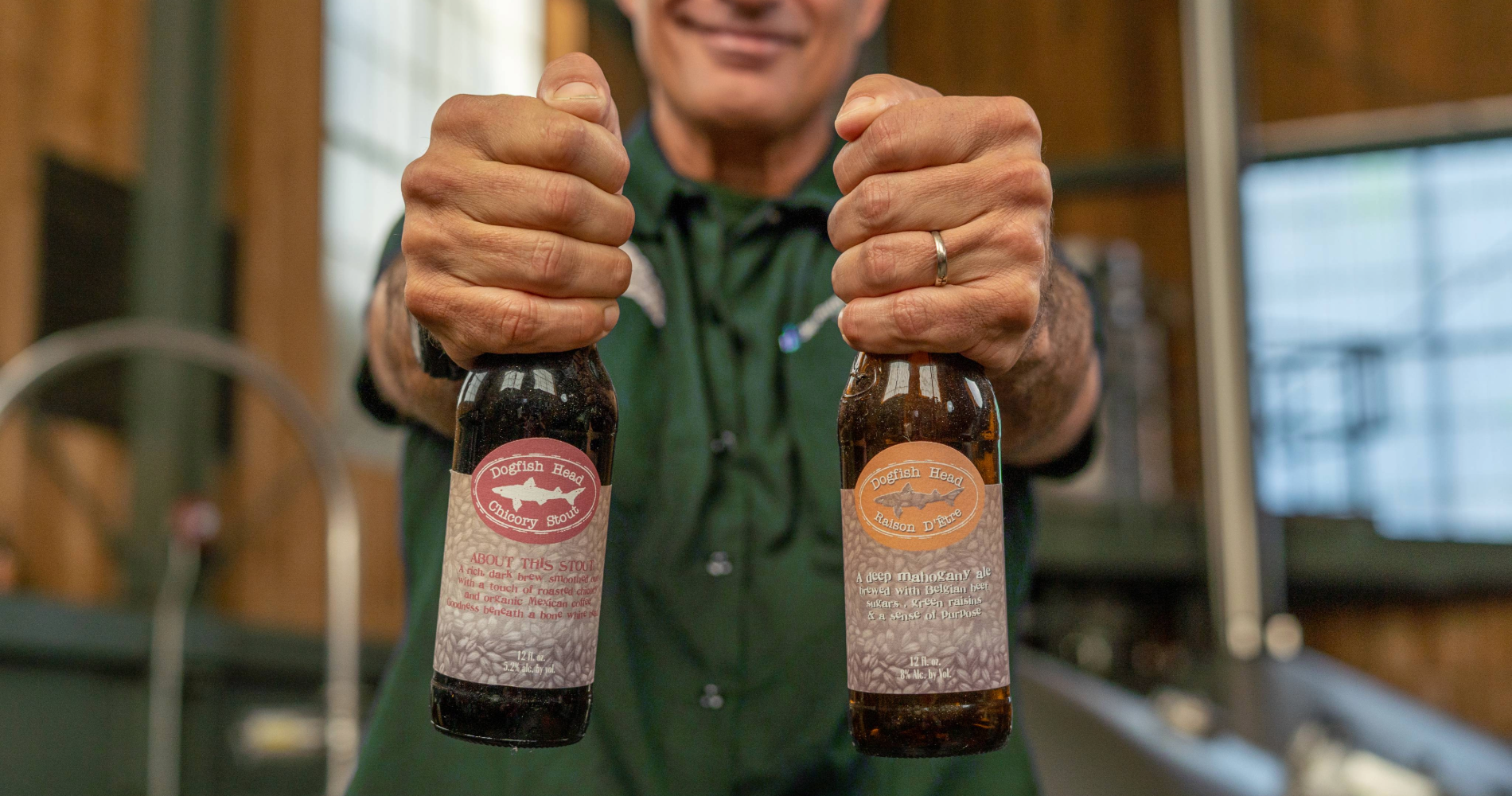 A photo of Sam Calagione holding bottles of Dogfish Head Chicory Stout and Raison D'Etre in their brewhouse in Milton