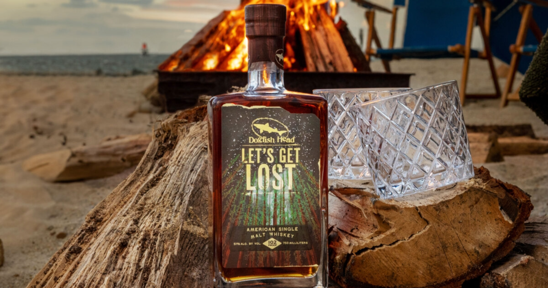 Bottle of Dogfish Head Distilling Co. on a beach with a bonfire behind it and the ocean