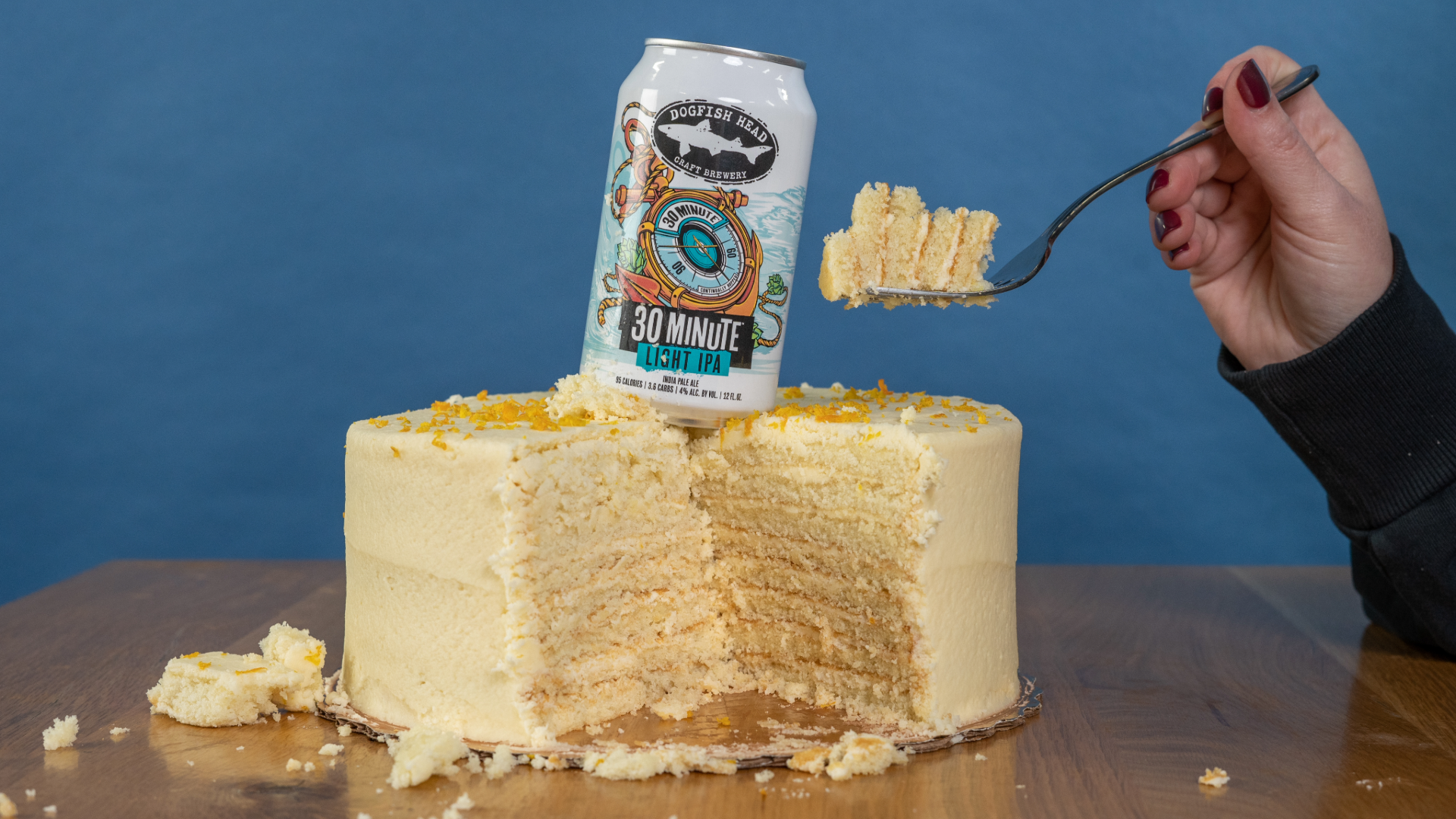 A Smith Island Cake with a slice missing and a hand holding a fork over the cake with a can of Dogfish Head 30 Minute Light IPA on top of the cake