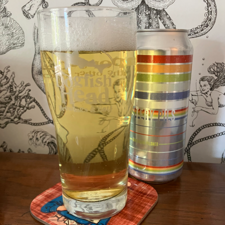 A pint of Mystery Pils, a light straw colored Pilsner, next to a 16 oz can of it on a bar with nautical wallpaper behind it