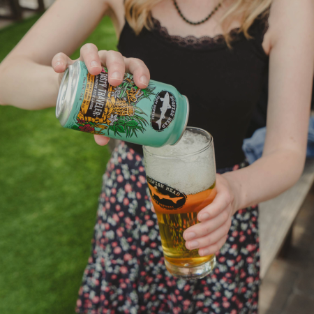 Someone pouring a Can of Tasty Traveler Lager with an illustration of bee surrounded by grass holding a honey comb