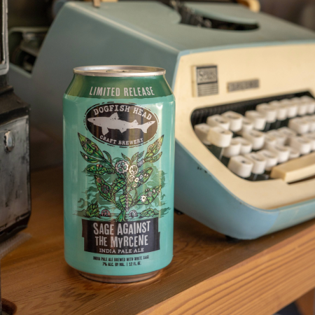 12 oz can of Dogfish Head Sage Against The Myrcene in a green can with cartoon sage artwork on a wooden desktop with a typewriter behind it