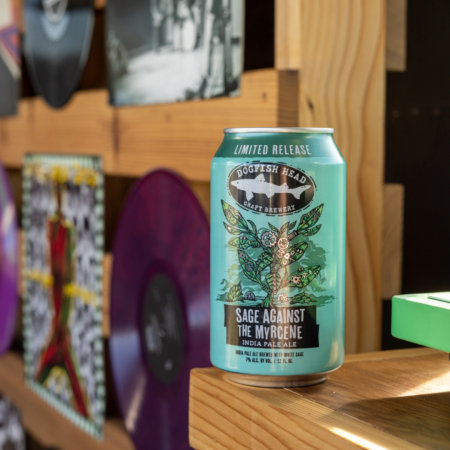 12 oz can of Dogfish Head Sage Against The Myrcene in a green can with cartoon sage artwork on a wooden desktop with a variety pack of beers behind it