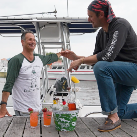 Sam Calagione and brad Leone with two pink cocktails in hurricane glasses on a dock in Lewes, Delaware with the S.S. Dogfish boat in the water behind them