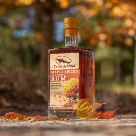 A bottle of Dogfish Head Maple Spiced Rum on a wooden deck with yellow and orange leaves and trees in the background