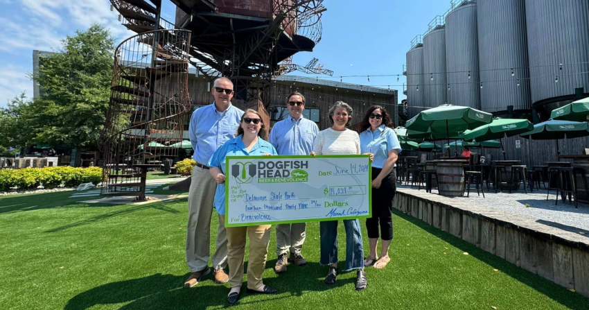 Staff from Delaware State Parks posing with a large check and Dogfish Head coworkers, including Mariah Calagione, in front of the Milton brewery