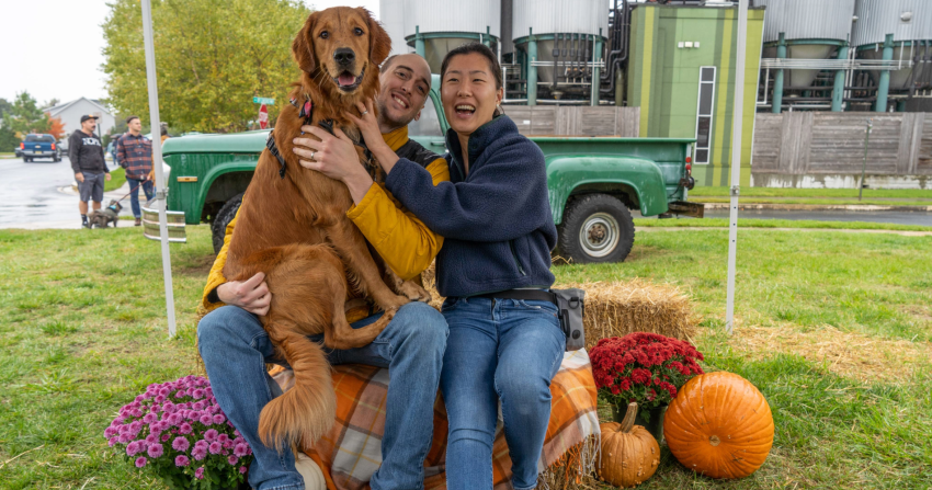 Couple posing with their golden retriever on a haybale in front of Dogfish Head brewery for Punkin Ale Fest