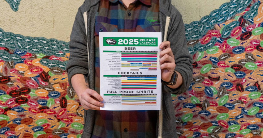 Photo of a Dogfish Head coworker holding the 2025 beverage calendar in front of a wall of Dogfish Head beer stickers