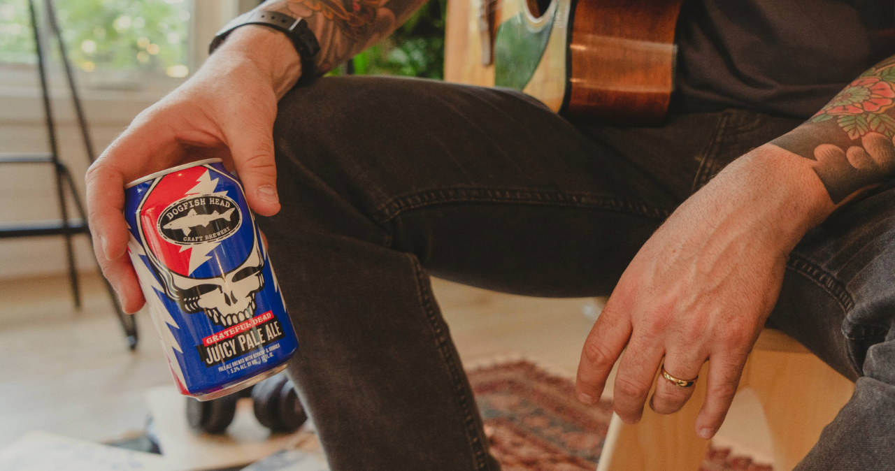 Dogfish Head x Grateful Dead Juicy Pale Ale 12 oz can in a person's hand who's sitting in a chair with a guitar on their lap