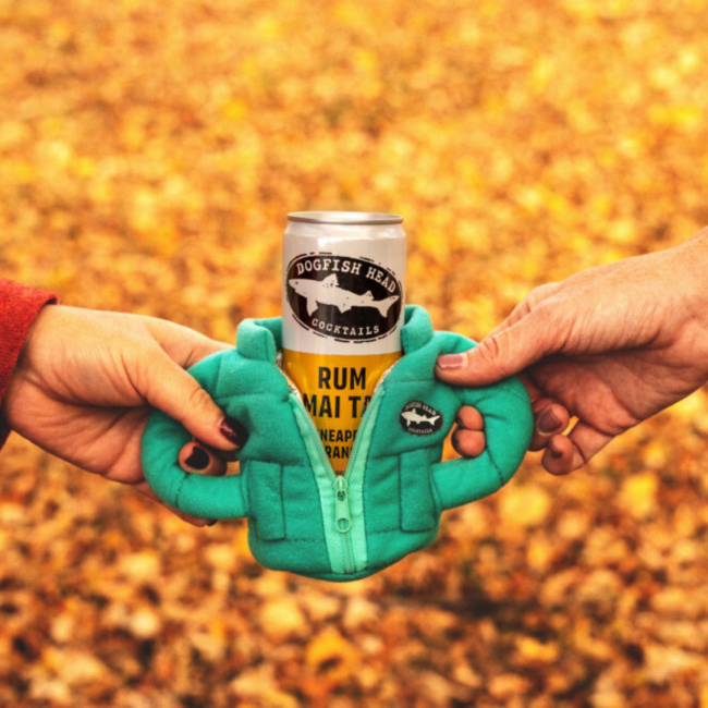 Pineapple Orange Rum Mai Tai in a puffer jacket can cooler being held on both sides by two hands with orange and yellow leaves on the ground in the background